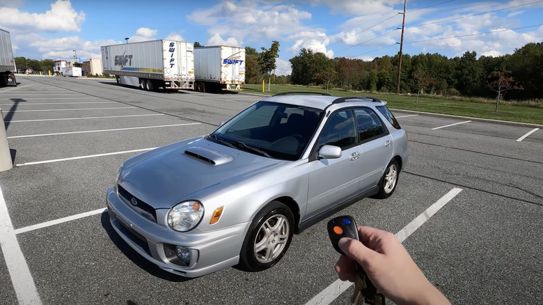 a silver subaru in a parking lot on a sunny day, several tractor-trailers in the background