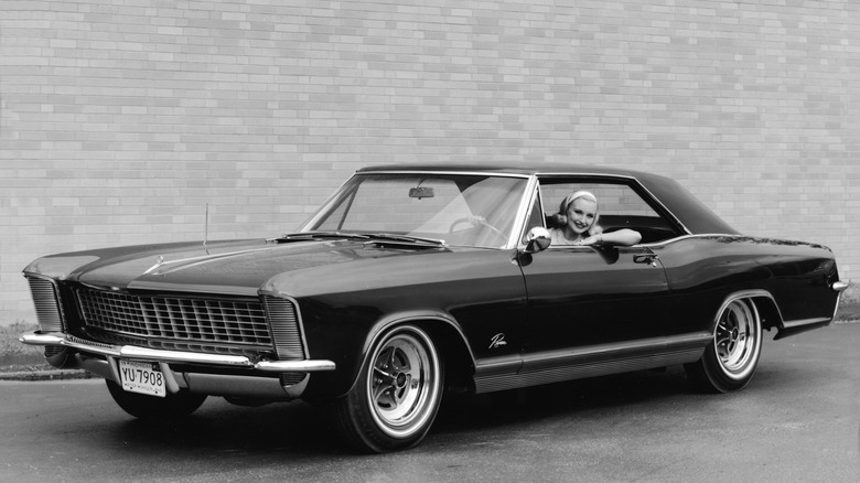 a black and white image of a 1965 buick riviera with a smiling woman in the driver's seat