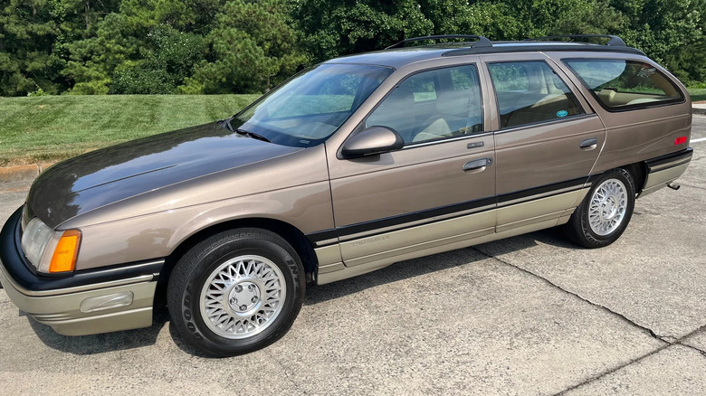 Side view of a copperish '87 Ford Taurus wagon parked on concrete in front of a lawn and hedges