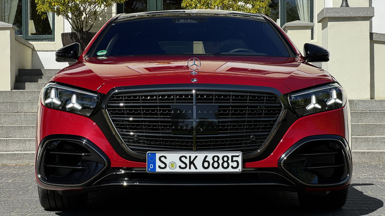 A head-on shot of a red 2027 Mercedes-Benz S580 parked in front of concrete stairs and a white building