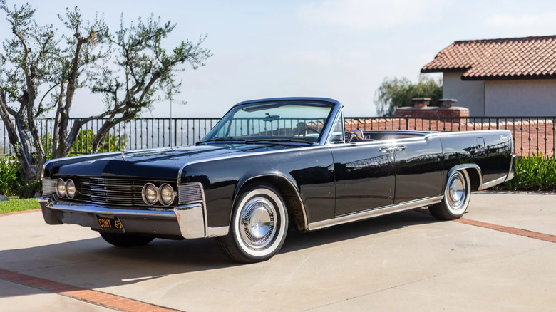 A black '65 Lincoln Continental Convertible parked on concrete in front of an iron fence a tree and a blue sky