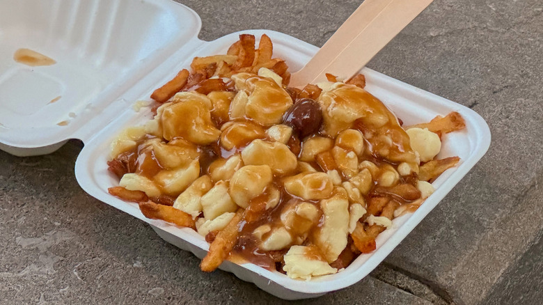 A serving of poutine successful a achromatic takeout container, featuring aureate fries topped pinch food curds and brownish gravy, placed connected a actual surface.