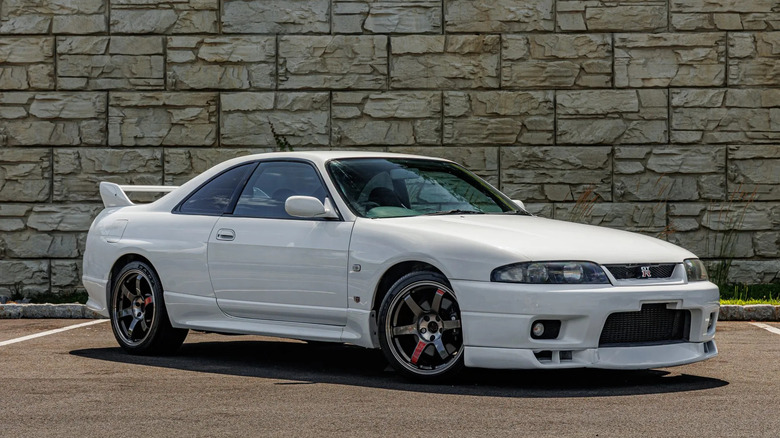 A white, 1995 Nissan Skyline GT-R (R33) in a parking lot with a large stone wall background.