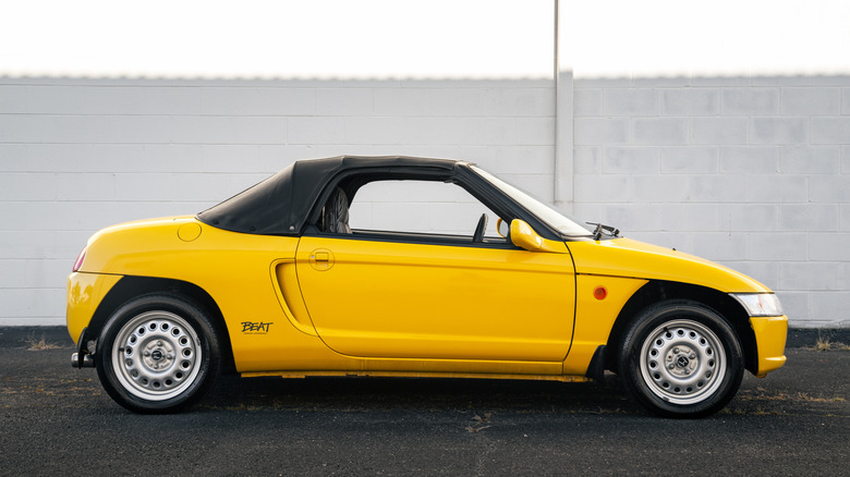 A yellow, Honda Beat with black cloth convertible top up, and steel wheels on a black top against a white brick building.