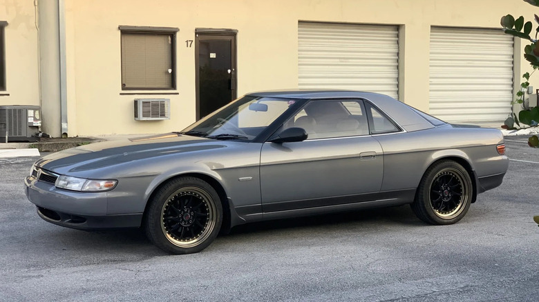 A driver's side shot of a grey 1991 Mazda Eunos Cosmo in front of a warehouse building with garage ports.