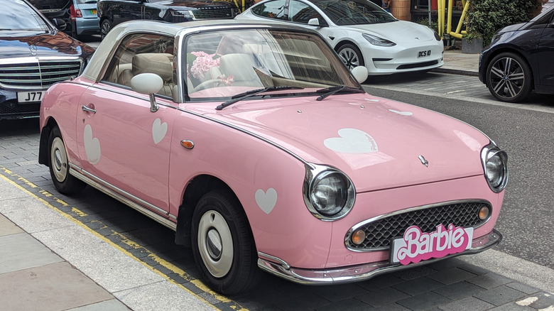 A pink Nissan Figaro with white heart decals and a barbie sign on the front license plate holder, parked along a city street.