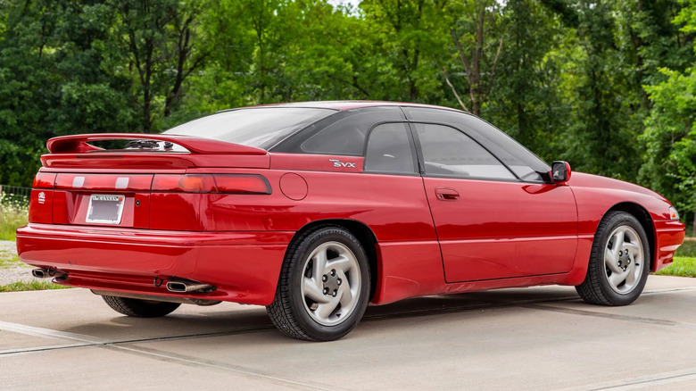 The rear passenger corner full-body shot of a red 1993 Subaru SVX on a cement driveway with trees in the background.