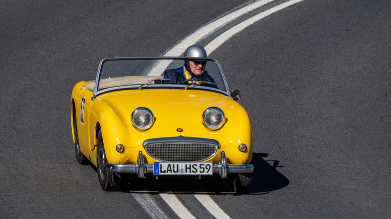 A yellow Bugeye Sprite driving down the road, a determined looking old man in a silver helmet at the wheel