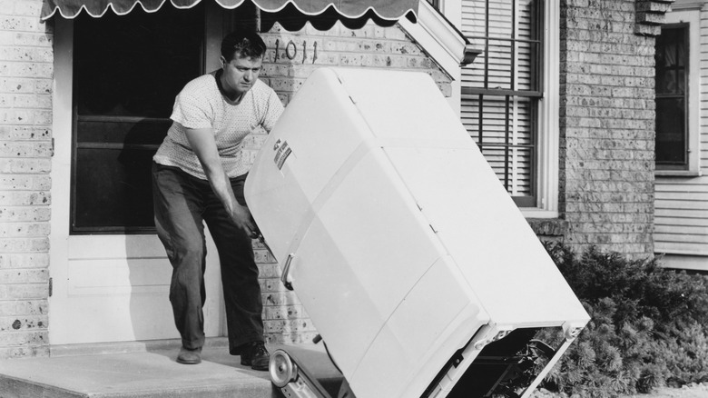 A black and white image of a man hauling a GM Frigidare up the front steps to his house.