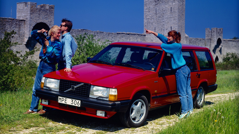 A red Volvo 740 Turbo wagon in front of a castle wall with three people leaning against it