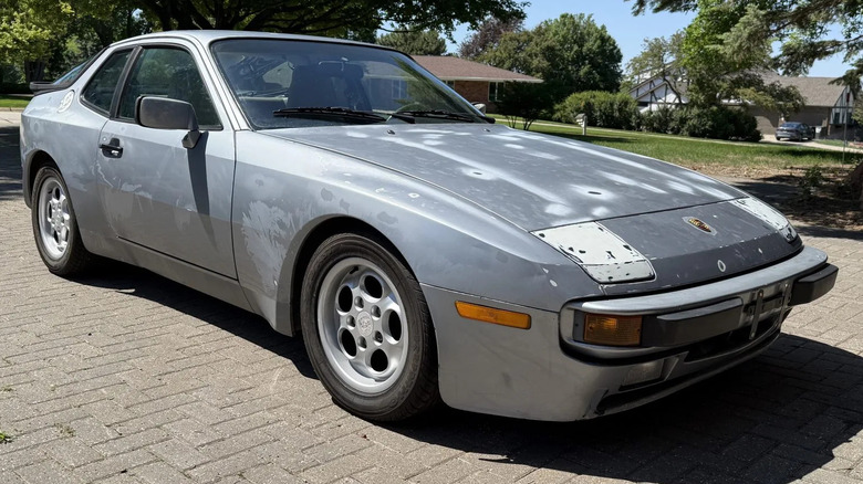 A front three quarters shot of a mostly silver Porsche 944 project car with bad paint and bondo on the headlights parked on brick in front of grass