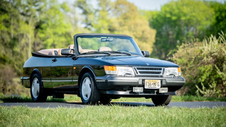 Front three quarters shot of a black Saab 900 Turbo convertible parked with its top down surrounded by grass and trees