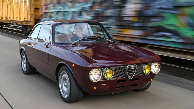 Front three-quarters rolling shot of a burgundy Alfa GTV driving past a graffitied train car