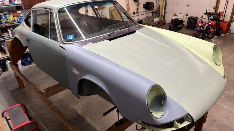 Front three quarters shot of a stripped down Porsche 911 T coupe body shell sitting on wood blocks in a garage