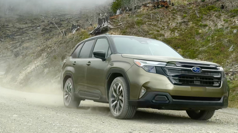 A front three quarters shot of a green Subaru Forester driving on a gravel road in front of a grassy hillside