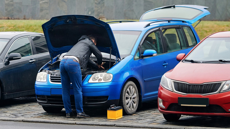 A person trying to fix a car with the hood up and battery on the ground.