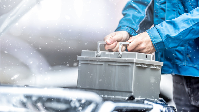 Two hands holding a car battery in snowy conditions.