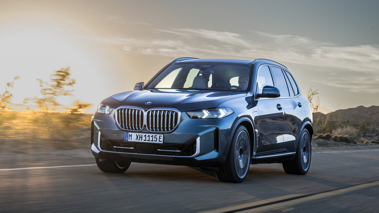 A 2024 BMW X5 xDrive50e in a front 3/4 view driving on a road with a setting sun in the background