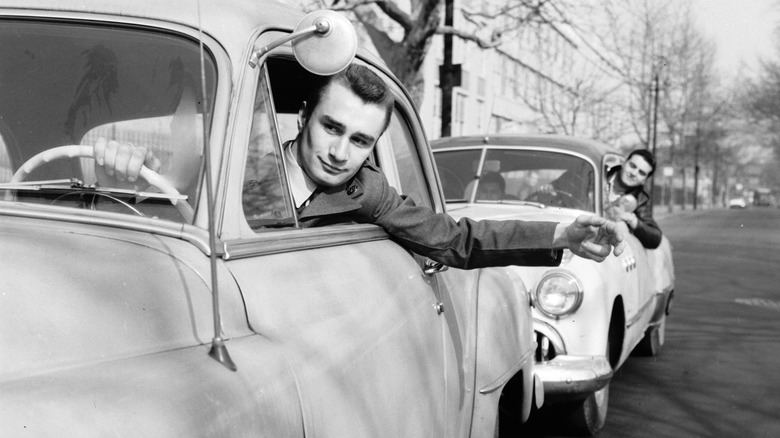 a man in a sedan hangs out his car window, a similar car is behind him with another man sticking his head out. The image is black and white, circa 1963
