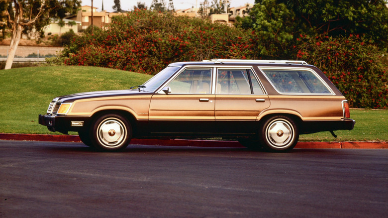 Ford Country Squire parked on black top in front of green hills and flowering bushes