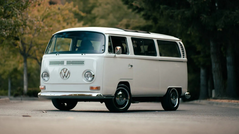 Front three quarters shot of a white '68 VW bus parked on asphalt in front of trees at sunset