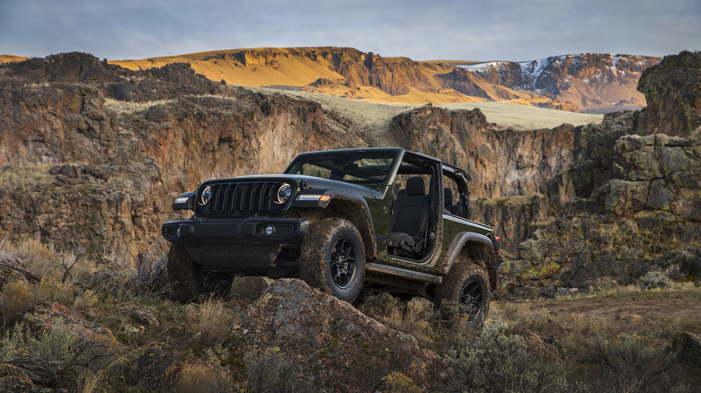 Front three quarters shot of a gray Wrangler 2-door with no doors or roof stopped while propped up on a rock in front of beautiful mountains