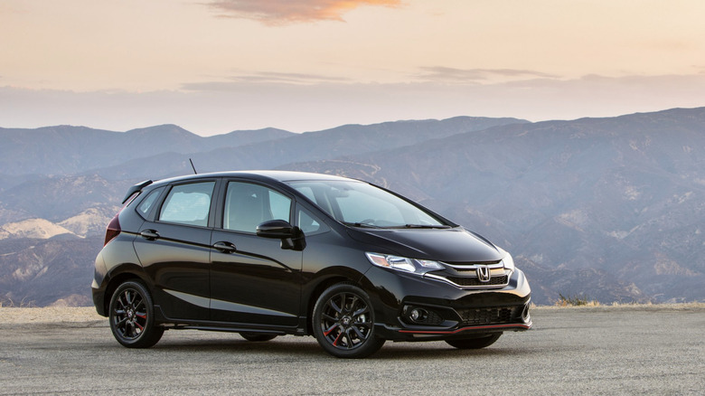 A black Honda Fit parked in a sandy lot with hills rolling into the background
