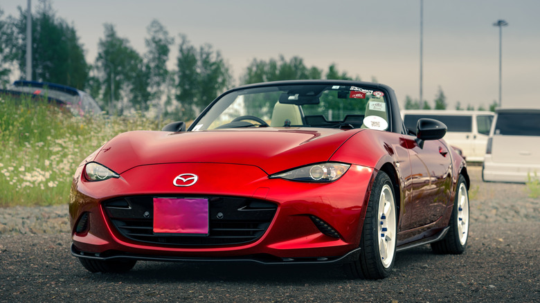 Igora Drive, Red Mazda MX-5 Miata convertible car parked on gravel with white vans and green trees in the background