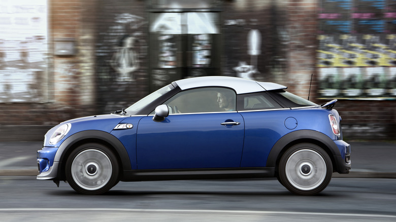 Side position of a bluish Mini Cooper S Coupe driving past a ceramic building pinch graffiti and posters connected it