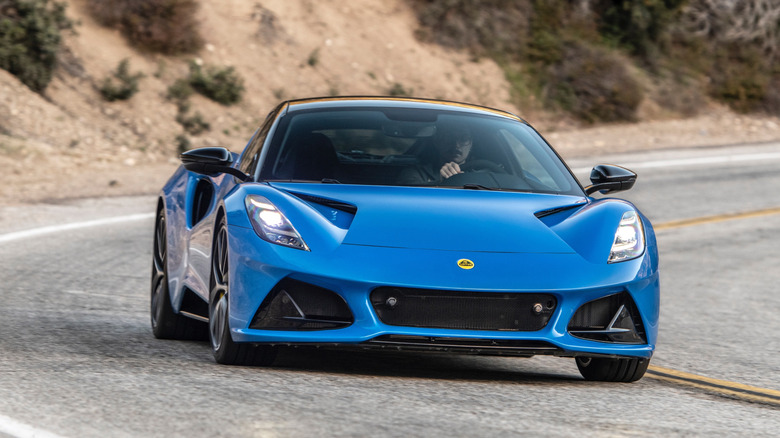 head-on shot of a blue Lotus Emira driving around a corner in front of a shrub-covered mountainside