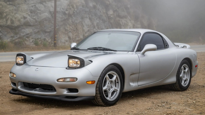 front three-quarters shot of a silver FD Mazda RX7 parked on dirt in front of a misty view of a rocky mountainside with its headlights on and popped up