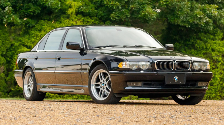 front three-quarters shot of a black E38 BMW 7 Series parked on gravel in front of bright green bushes