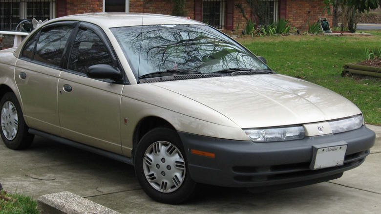 front three-quarters shot of a light gold Saturn SL1 with black plastic bumpers and hubcaps parke don concrete in front of grass and a brick home