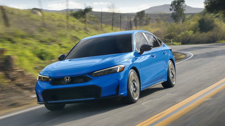 A blue Honda Civic hatchback driving around a corner on a mountain road with green hills and trees in the background
