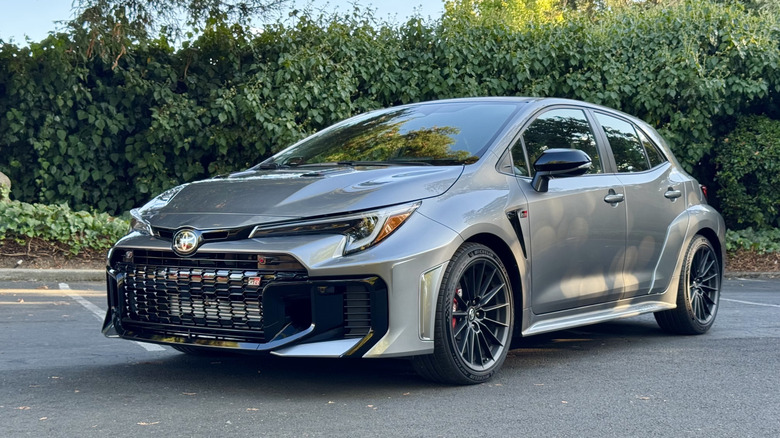 A front three quarters shot of a silver Toyota GR Corolla parked in a parking lot in front of a hedge