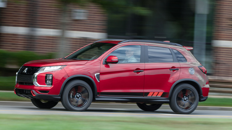 A side view of a red Mitsubishi Outlander Sport driving through a city in front of grass and brick buildings