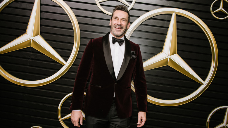 Actor Jon Hamm, wearing a velvet tuxedo, standing in front of a backdrop with Mercedes-Benz logos on it.