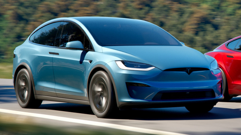 A front three quarters shot of a blue Tesla Model X driving on a road next to a red Model S with trees in the background