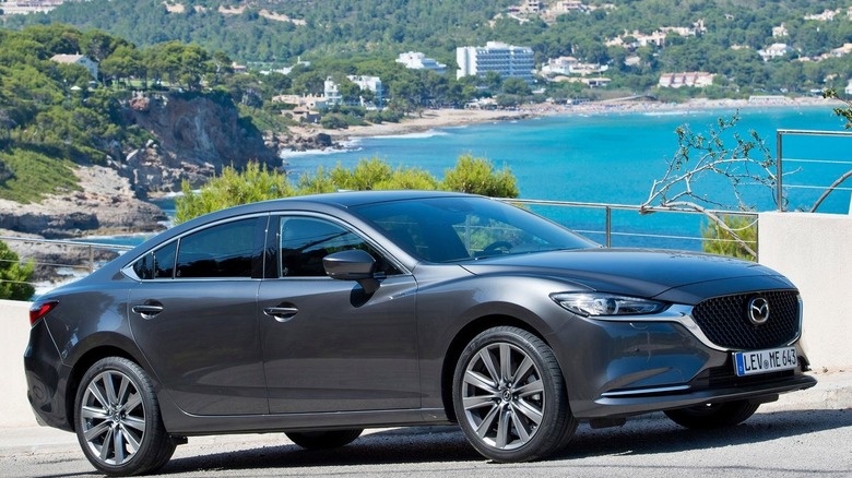 Front three quarters shot of a dark gray Mazda 6 parked on a cliffside road in front of a view of blue water and lots of trees