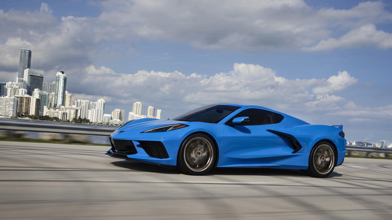 side view of a blue C8 corvette driving on a bridge in front of a cityscape and a cloudy sky