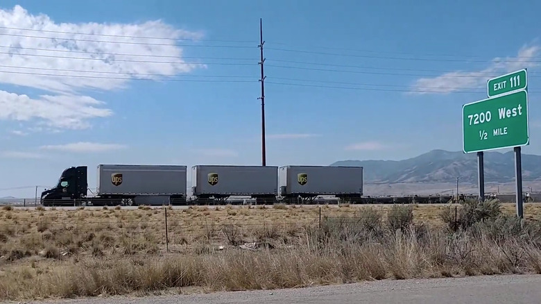 A UPS triple trailer semi truck being driven down a highway in a western state with mountains in the background