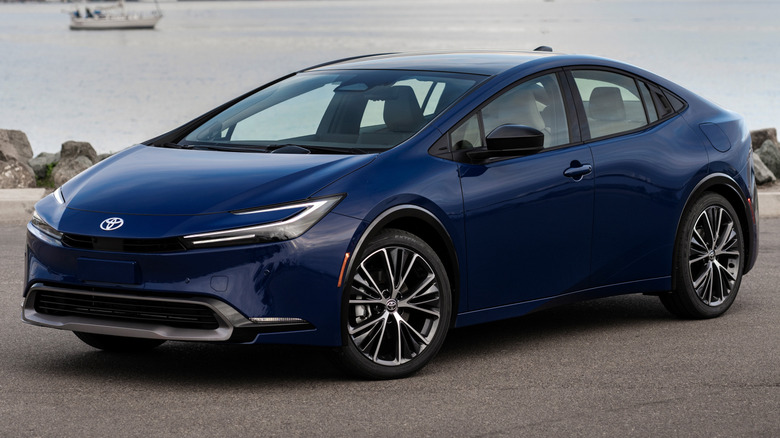 Reservoir Blue 2025 Toyota Prius parked on a paved surface with a body of water and city skyline in the background