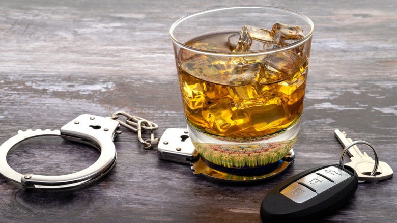 A glass of dark liquor sits on top of handcuffs and next to car keys