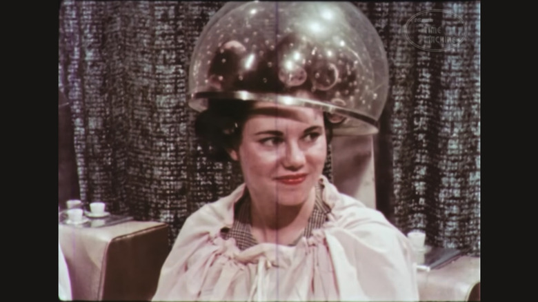 One of the flight attendants sits in a salon chair with a dryer bubble over her hair in curlers.