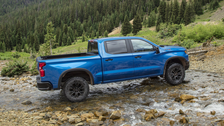 Side view of a blue Chevy Silverado ZR2 making a shallow river crossing with mountains and trees in the background