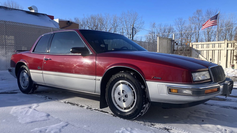 Front three quarters shot of the burgundy and silver Riviera parked on snow in front of a fence and an American flag