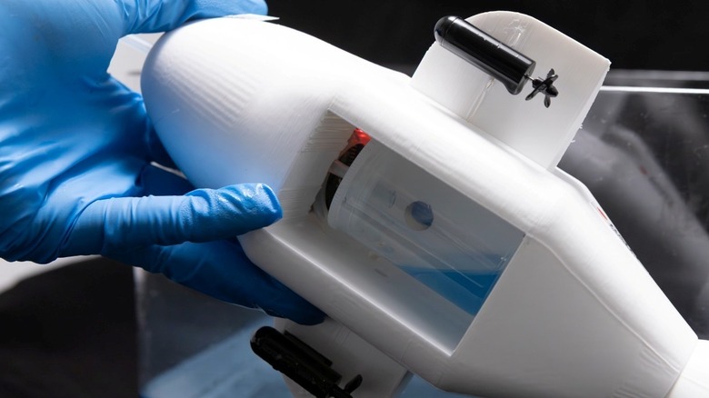A gloved hand holds the actual electronic dolphin device, to show the oil tank which contains a bit of an oil solution.