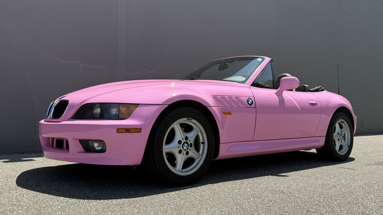 A very pink 1997 BMW Z3 roadster in a parking lot parked in front of a building painted in dark grey brown color.