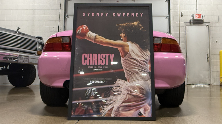 The poster from the Christy film signed by Sydney Sweeney, Christy Martin, the film's director and one of the cast members leans against the back end of a pink convertible BMW in a garage setting.