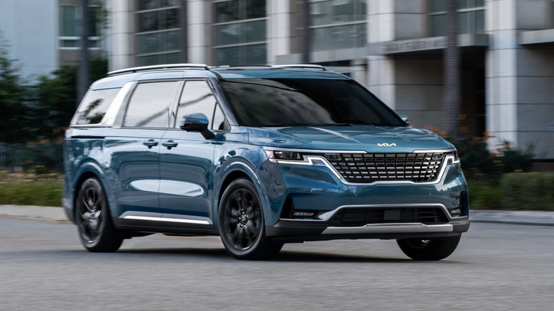 A blue 2022 Kia Carnival being driven in front of building.
